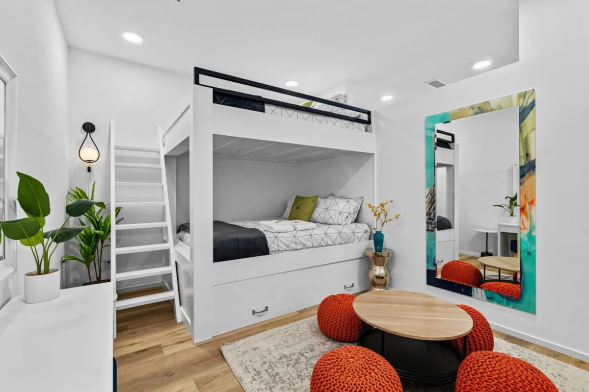 A bright, inviting loft space featuring a lofted bed, colorful mirror, round seating area, and lush green plants. The setup feels airy and playful, with natural light filtering through, offering hints of socializing and relaxation. The seating area is perfect for morning coffee or casual work sessions, while the overall space suggests a flexible environment ideal for both productivity and downtime.
