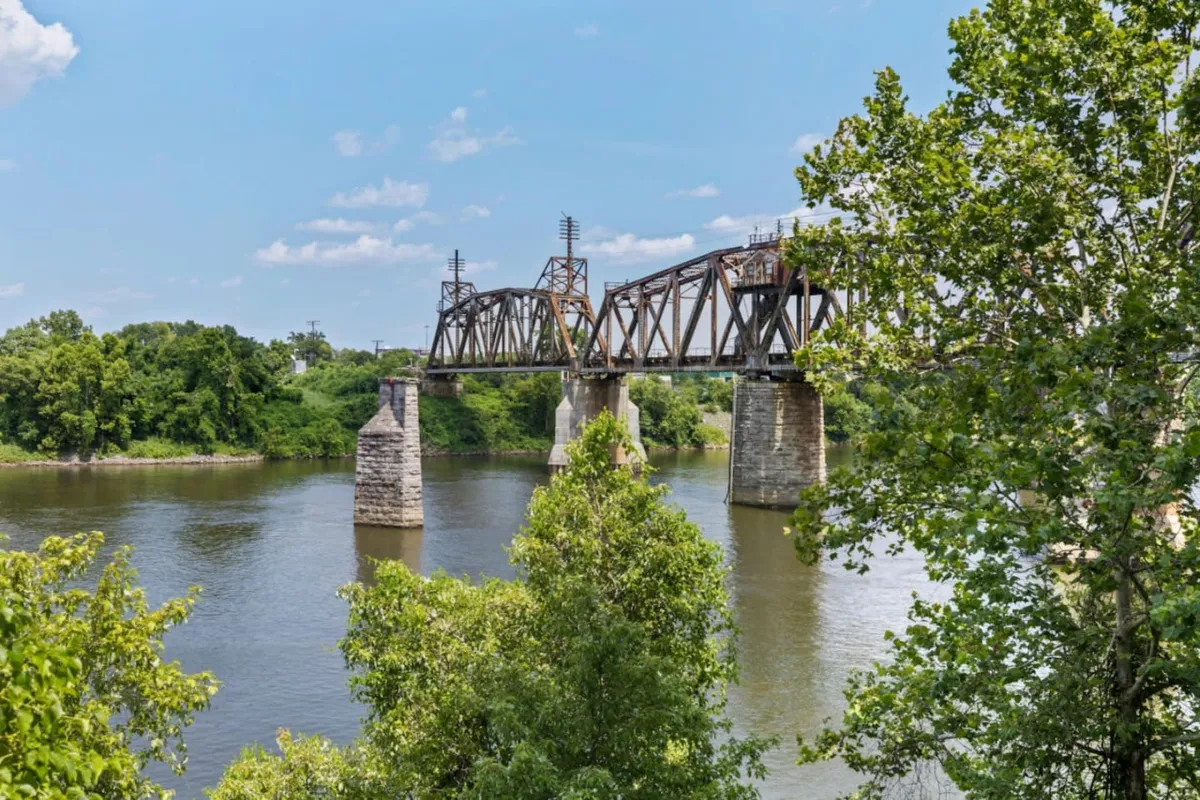 The image showcases a tranquil river at sunset, bordered by lush greenery and a striking rusted train bridge. The serene water reflects the soft colors of the sky, suggesting an atmosphere perfect for relaxing, observing wildlife, or enjoying a peaceful nature walk.