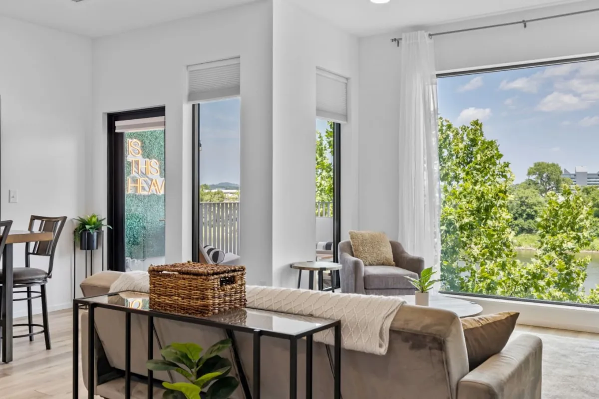 A bright, modern living space with a large window offering a scenic view, a comfortable seating area, and a stylish coffee table. Soft natural light floods the room, suggesting a connection to the outdoors and the city beyond.