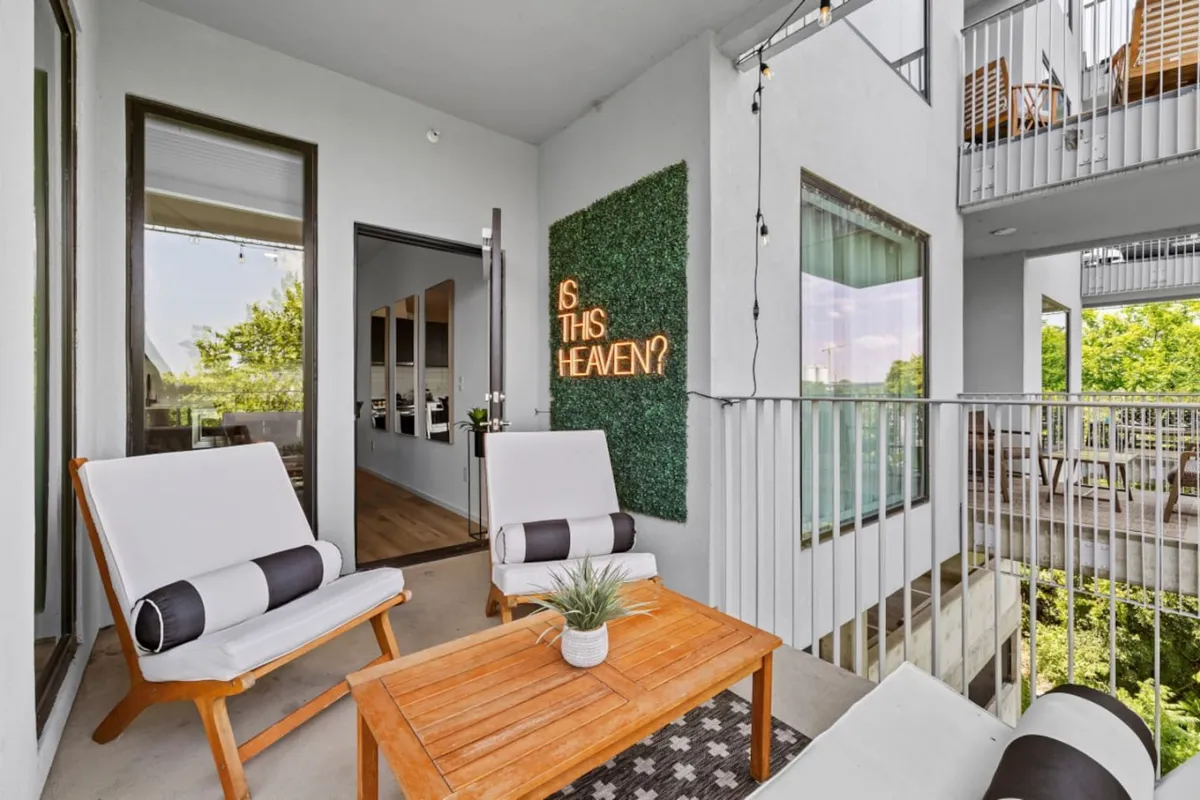 An inviting outdoor lounge area featuring a lush green wall with playful 'IS THIS HEAVEN?' text art, complemented by comfortable lounge chairs and a wooden coffee table. String lights add a festive touch, and the space feels perfect for selfies or pre-night-out relaxation. The area is bathed in natural light, offering a stylish, Instagram-worthy backdrop.