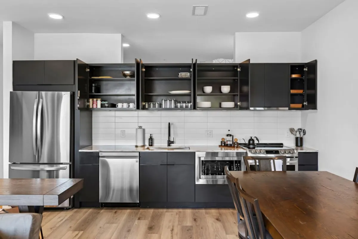 A contemporary kitchen and dining setup with stainless steel appliances, open shelving, and a large wooden dining table. The space feels lively and ready for group gatherings, making it ideal for shared meals or celebrations.