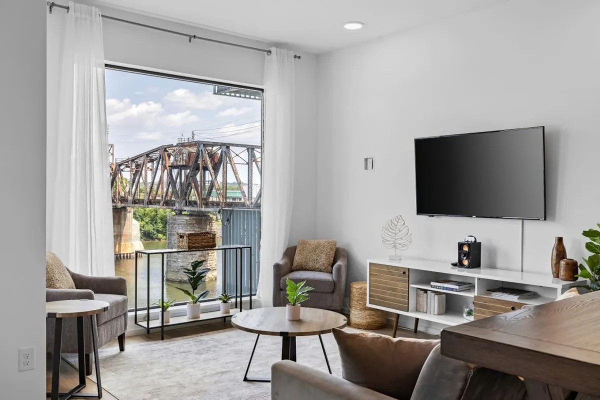 A cozy and contemporary living area with large windows offering a clear view of the bridge and river beyond. The space is furnished with comfortable armchairs, a modern television, and a stylish coffee table, inviting guests to relax, entertain, and enjoy the scenery.
