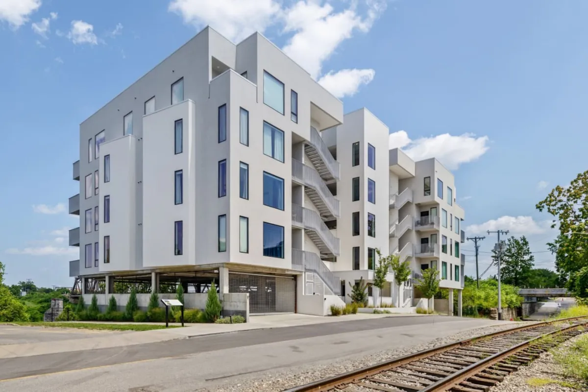 A contemporary exterior view of the apartment building, showcasing modern architecture with expansive windows and inviting balconies. The scene hints at community life, evening strolls, and proximity to the city's energy, with sunny weather highlighting the building's appeal.