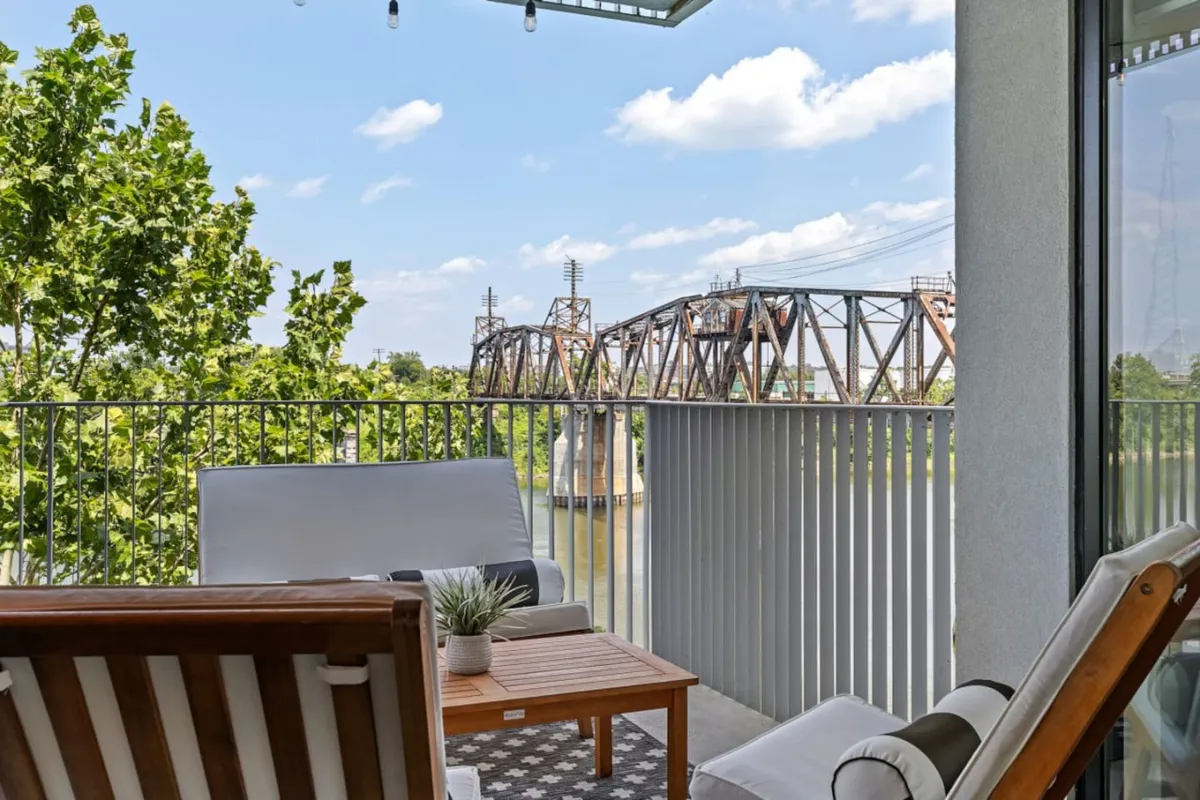 A tranquil outdoor seating area with a view of the bridge and lush green foliage. The space feels inviting for relaxing or social gatherings and subtly hints at the nearby train tracks, connecting to the city soundtrack mentioned in the narrative.