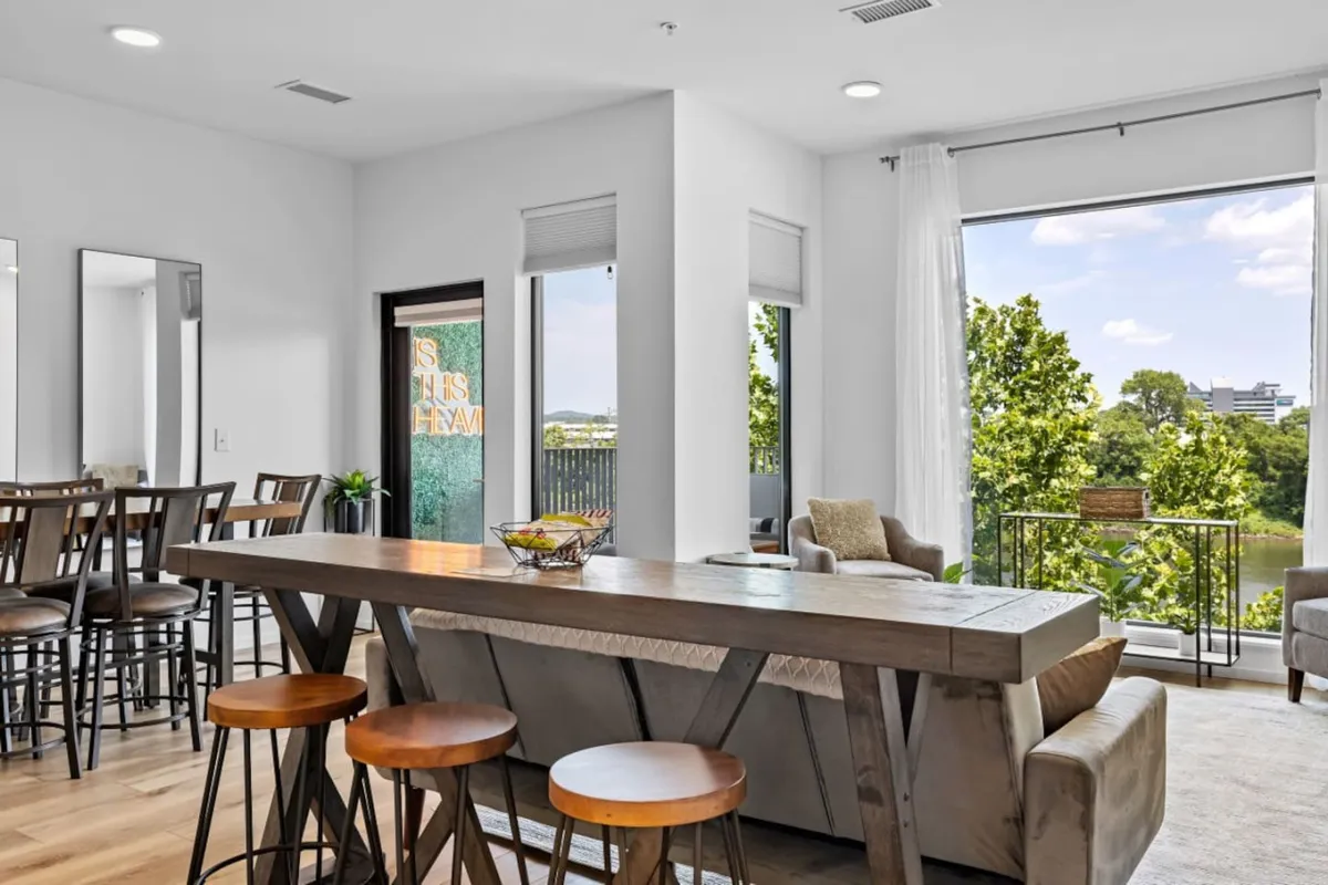 A bright, inviting interior scene featuring a large wooden dining table, a comfortable sofa for lounging, and expansive floor-to-ceiling windows that flood the space with natural light and scenic vistas.