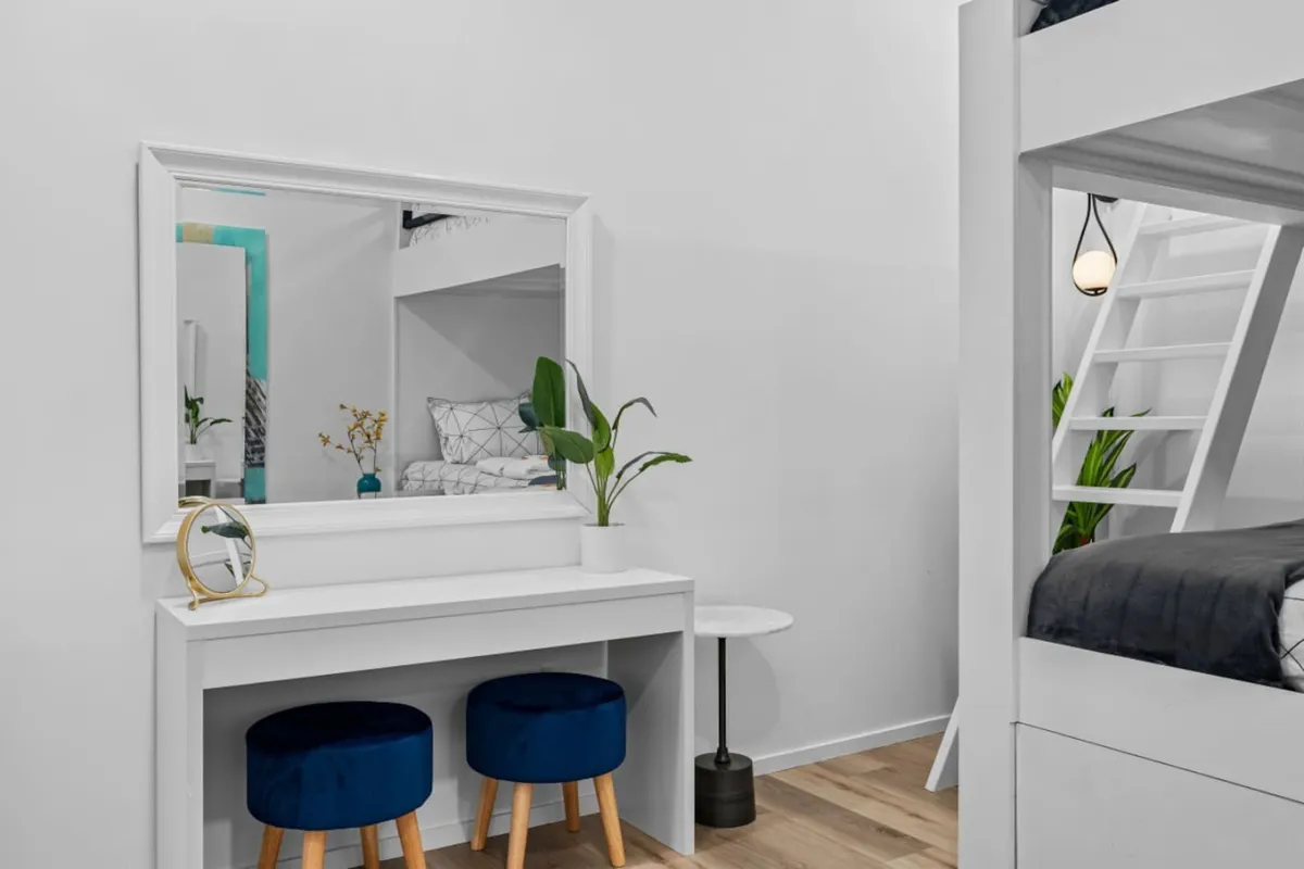 A bright bedroom scene with a bunk bed and a single bed, adorned with a mirror and vanity table, blue stools, and a touch of greenery. The space feels youthful and functional, ideal for families or groups.