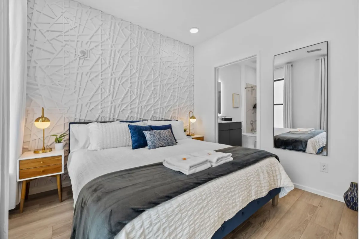 A serene bedroom featuring a plush king-sized bed dressed with decorative pillows, flanked by nightstands with lamps. The backdrop is a textured accent wall, and the mood is quiet and cozy, inviting relaxation and restful sleep.