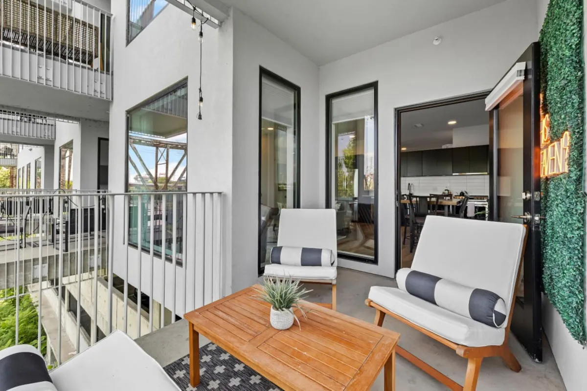 A private balcony furnished with white lounge chairs and a wooden coffee table, framed by lush greenery. The setting is ideal for relaxing, entertaining, and enjoying the outdoors with views of the urban landscape.