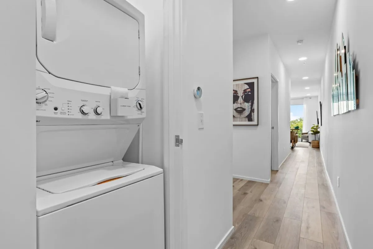 An efficient laundry area with a stacked washer and dryer set in a hallway filled with natural light. A piece of framed artwork adds a welcoming touch to the practical space.
